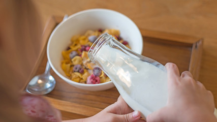 kid hand pouring fresh milk on the white cup, cornflakes cereal, strawberry, blackberry and milk in a white bowl, selective focus