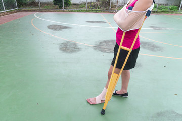 Injured woman wearing sportswear painful arm with gauze bandage, arm sling and wooden crutches on green floor.