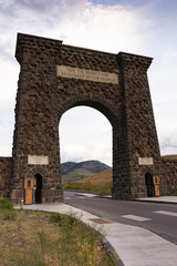 The Roosevelt Arch at the entrance of Yellowstone National Park with its engraved with "for the benefit and enjoyment of the people". Cloudy sky is in the background and a road in the foreground.