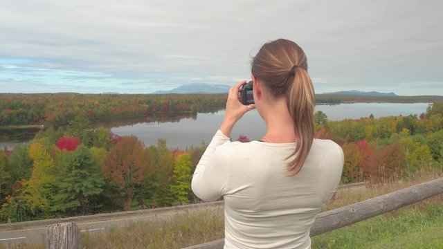 Young brunette traveler taking photos of breathtaking nature in autumn. Girl tourist photographing stunning lake surrounded by colorful autumn forest. Female explorer on leaf-peeping trip on viewpoint