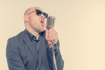 Unshaven bald man in a gray suit and sunglasses holding a microphone and singing. Toned