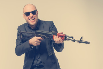 Fototapeta na wymiar Smiling bald man in a gray suit and sunglasses holding a machine gun in his hands. Toned