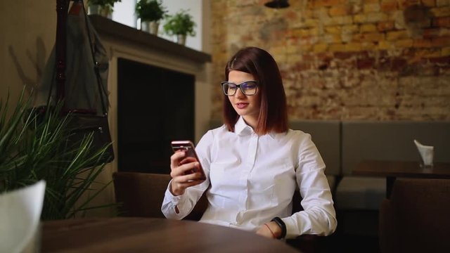 Charming woman with beautiful smile reading good news on mobile phone during rest in coffee shop, happy Caucasian female watching her photo on cell telephone while relaxing in cafe during free time.