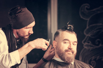 Hairdresser in a black cap and a black apron cuts with hairclipper a man with a clip on the head