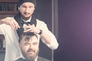 Stylist in black hat, bow tie and scarf makes hair a man with a beard