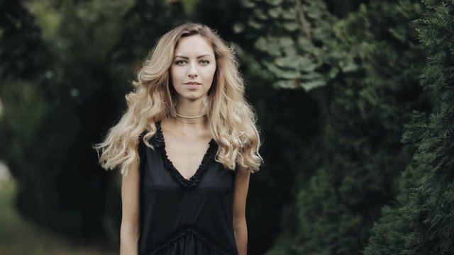 Beautiful woman with curled hair and earrings walking in city green park, slowmotion