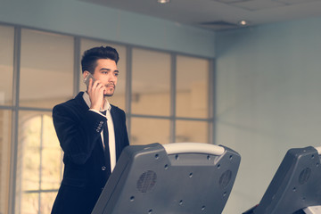 Handsome young man in a black suit, white shirt and tie talking on the phone on a treadmill in the...