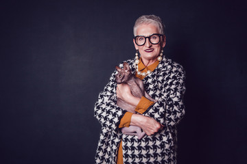 Stylish old woman in glasses holds a Sphynx cat on a black background.