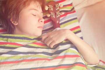 Obraz na płótnie Canvas Funny girl teenager in the gray pajamas lying in bed with ukulele in his hand on colored striped pillow. Toned