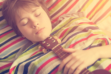 Funny girl teenager in the gray pajamas lying in bed with ukulele in his hand on colored striped pillow. Toned