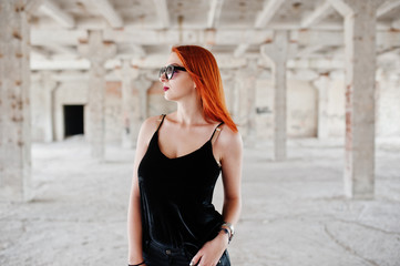 Red haired stylish girl in sunglasses wear in black, against abadoned place.