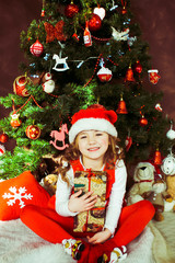 Little girl in red dress sits with a present box before a Christmas tree
