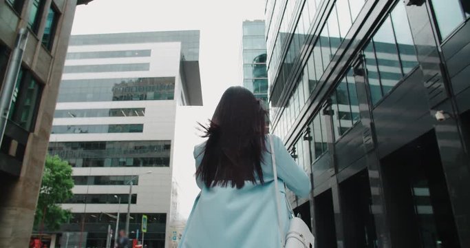epic Steadicam shot of a young beautiful Asian girl with smartphone going to work around office buildings, back view, flowing hair