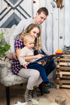 Portrait of young family: mom, dad and little son. All of them sitting in the decorated room. Mom holding a book and showing something to her little son. Concept of Merry Christmas and New Year