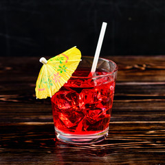 Top view of glass of red cocktail with ice cubes and straw umbrella on dark wooden table, close-up