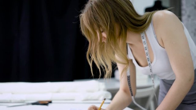 Close up of young woman with tape-line on her neck standing bending over the table in tailoring studio sketching future dresses. Designer creating new fashion collection doing outline with pencil.