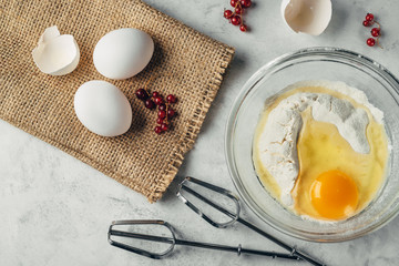Top view photo of a broken egg on flour in a glass plate. Mixer fits on a white surface. Eggs and currant on a brown bagging. Ingredients for healthy rustic breakfast.