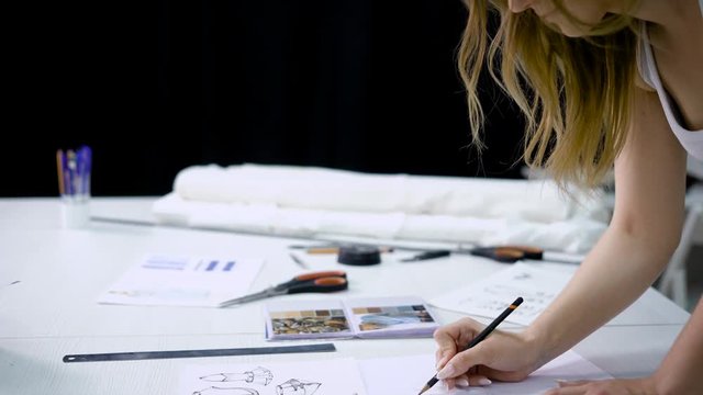 Close up of young fair-haired woman standing bending over working table in tailoring studio and drawing future dresses. Clothes designer is creating new fashion collection doing sketches with pencil.