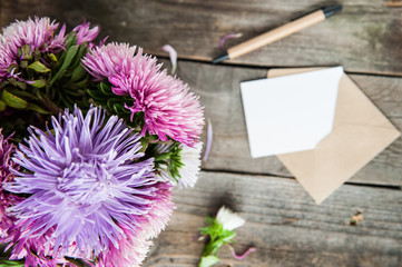 Top view Multicolor aster flowers bouquet, pen, blank white greeting card and craft paper envelope on rustic wooden table.. Postcard mock up. Autumn flowers. Selective focus. Space for text. Flat lay
