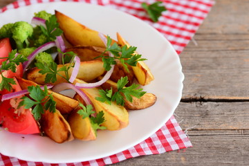 Roasted potato wedges, cooked broccoli, fresh tomato slices with spices, red onion, parsley leaves on a white plate and on an old wooden background. Easy vegetable dish