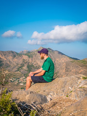 Man with beard and cap sits at the top of a mountain and looking out on the view.