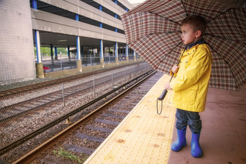 boy at the railway station in rain. cute kid in a yellow raincoat under an umbrella outside. The concept of commute. Copy space for your text