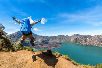 Hiker man with backpack spreading hand, jumping and happy with active volcano Baru Jari, Lake Segara Anak and summit of Rinjani mountain after finished climbing at Rinjani mountain, Lombok, Indonesia.