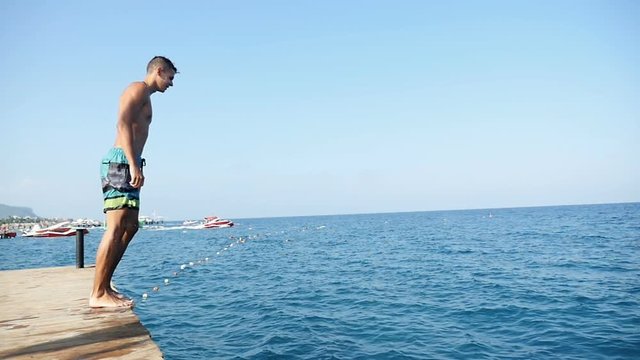 A young sportive man jumps straight from a pier into the sparkling sea waters of a Turkey resort in summer on a sunny day in slow motion. He is shot in profile with nice skyscape