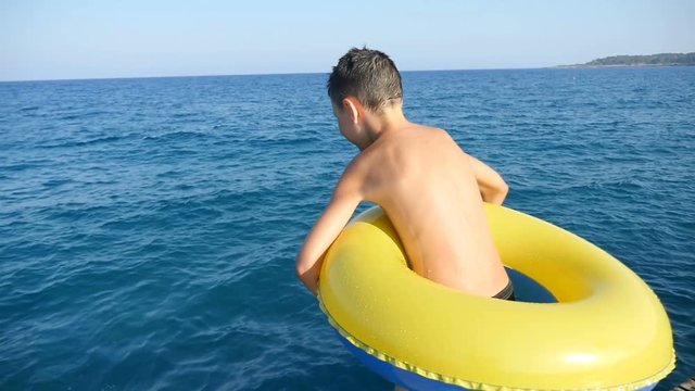 A seven-year-old boy with a yellow and blue inflatable ring jumps into the Mediterranean sea from a pier in summer in slow motion. The horizon line looks great