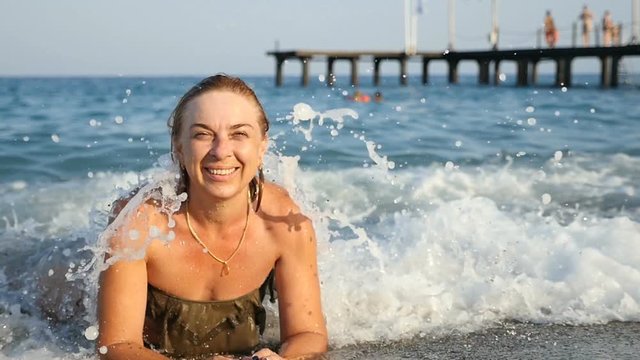 A jolly young woman smiles, frolics, and lies in the rough blue waters of the Mediterranean sea coast on a sunny day in summer in slow motion. She looks excited and lucky