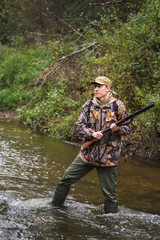 Hunter with a backpack and a hunting gun in the autumn forest. The man is on the hunt. The hunter crosses the forest river.
