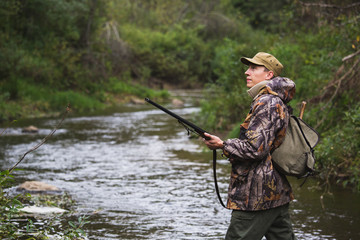 Hunter with a backpack and a hunting gun in the autumn forest. The man is on the hunt. The hunter crosses the forest river.