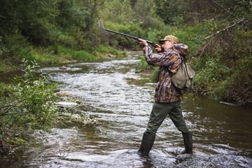 Naklejka premium Hunter with a backpack and a hunting gun in the autumn forest. The man is on the hunt. The hunter crosses the forest river.