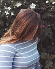 A very beautiful and cheerful girl stands on the street near a bush with white flowers.