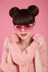 Fashion style teen girl model in knitted sweater. Brunette in heart sunglasses with matte lips and bun hairstyle posing over studio pink background.