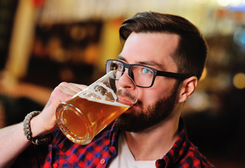 Cute young man in a plaid shirt drinking beer sitting on a chair in a pub