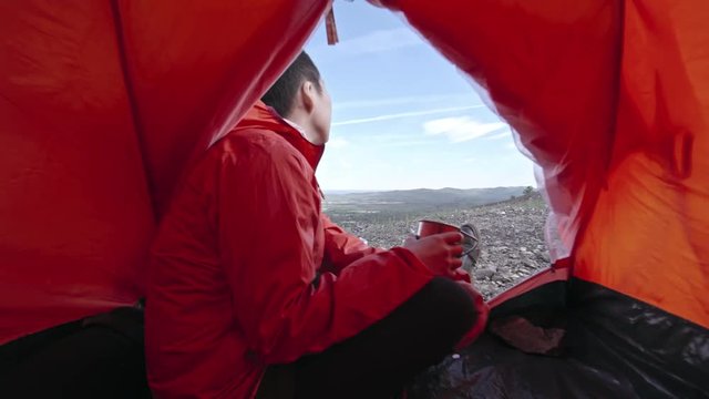 View from inside of camping tent of young Asian woman with short haircut drinking tea from metal mug