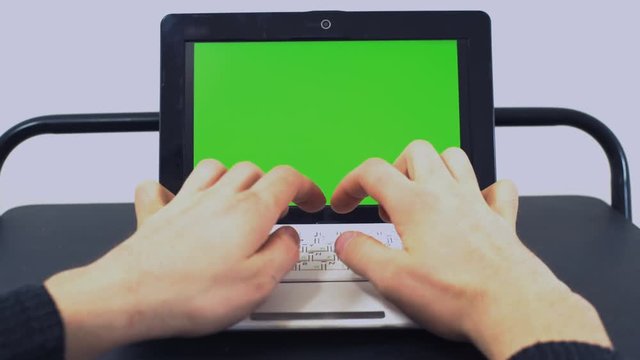 Male (Male hands) - The office worker sits in the office behind the swine in a robust place and prints on the keyboard of a white laptop with a green screen on a light background of the interior.