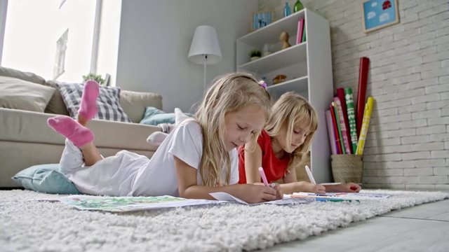 Ground level shot of cute little girls with blond hair lying on carpet in airy living room and laughing while drawing pictures with felt-tip pens