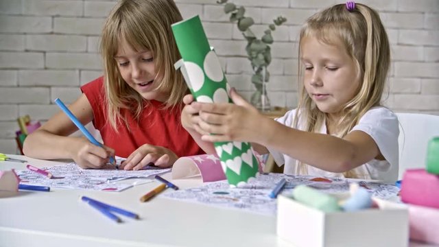 Cute girl with blond hair sitting before table and coloring with pencils while her little sister choosing stickers for her picture
