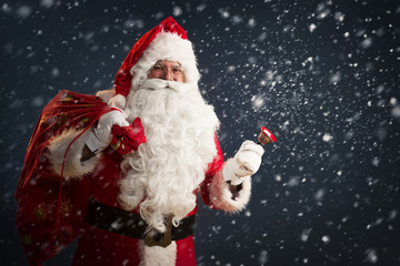 Santa Claus holding a bag with presents and ringing a bell on a dark background with snow 