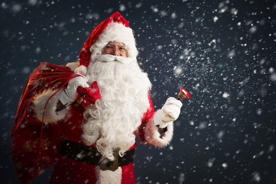 Santa Claus Holding A Bag With Presents And Ringing A Bell On A Dark Background With Snow 