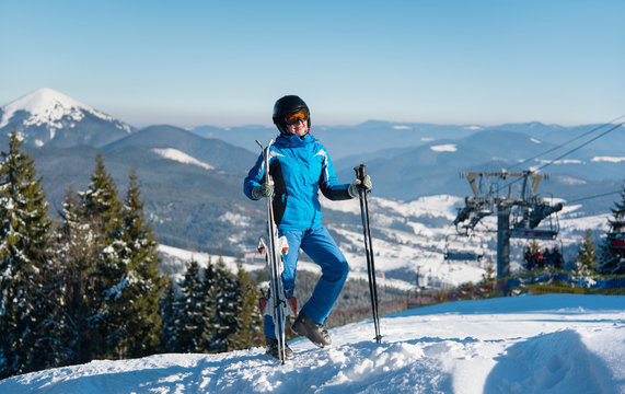 Full length shot of a happy female standing with her skis on top of the mountain at ski resort, smiling joyfully, enjoying winter sunny day. Blue sky, mountains, forests, ski lift on the background