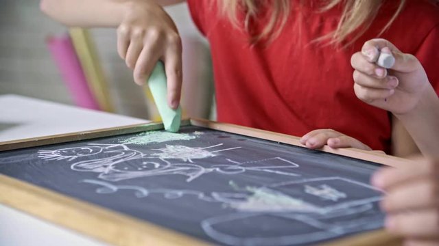 Mid-section of unrecognizable little girls drawing family and house with chalk on blackboard