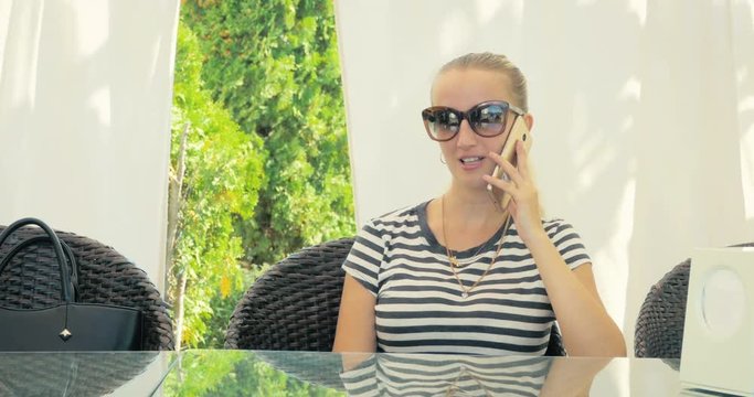Young beautiful blonde in sunglasses talking on the smart-phone sitting on a chair in summer cafe.