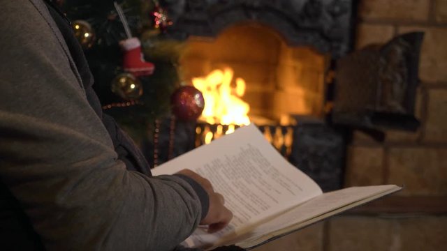 Senior man reading book near the fireplace and Christmas tree at xmas eve