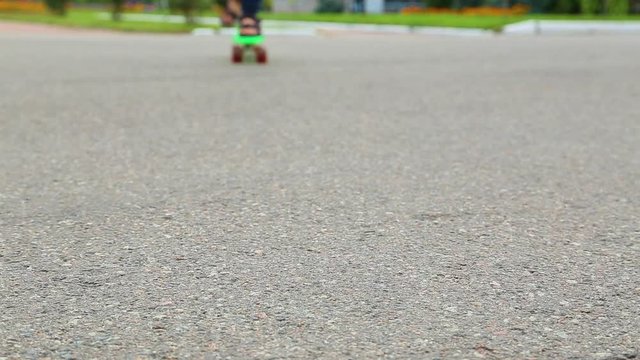 Closeup of male feet of child skating on green plastic penny-board on road in city. White kid of 10 years has fun in city park on warm summer or autumn day. Real time full hd video footage.