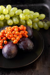 Fototapeta premium Green grapes, blue plums and orange rowan berries on a black plate for designer in retro style