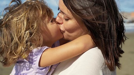 Woman kissing little blond daughter smiling in front of the ocean looking camera slow motion - Powered by Adobe