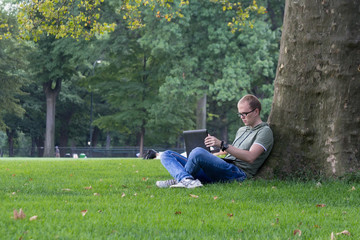 Young man in glasses work on laptop sitting on the grass near the tree in park. Maybe student or hacker